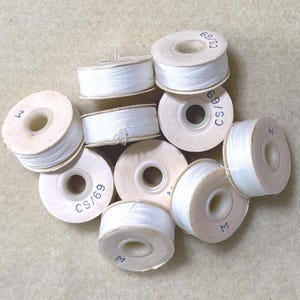May include: A group of ten white thread spools with brown cardboard bobbins. The bobbins have various markings, including "CS/69", "M", and "69/CS".