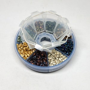 Puede incluir: Un recipiente de almacenamiento de cuentas de plástico transparente con tapa. El recipiente tiene siete compartimentos, cada uno lleno de un color diferente de cuentas de semillas. Las cuentas son en tonos de oro, marrón, blanco, verde y azul.