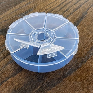 May include: A clear plastic rotating pill box with seven compartments. The lid is open and the compartments are visible.