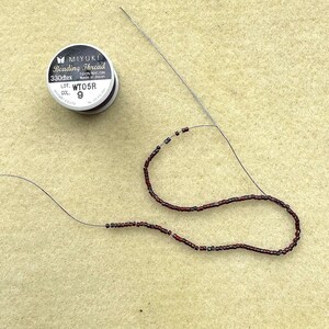 May include: A spool of Miyuki beading thread with the text "Beading Thread" and "330dtex". A needle is threaded with dark red and black beads. The beads are strung on a thin, dark thread, forming a curved line.
