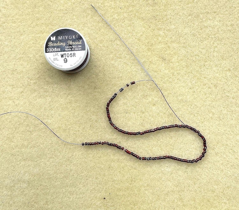 May include: A spool of Miyuki beading thread with the text "Beading Thread" and "330dtex". A needle is threaded with dark red and black beads. The beads are strung on a thin, dark thread, forming a curved line.