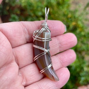 May include: A silver wire wrapped agate pendant in the shape of a tooth. The agate is brown and white with a natural, organic shape.