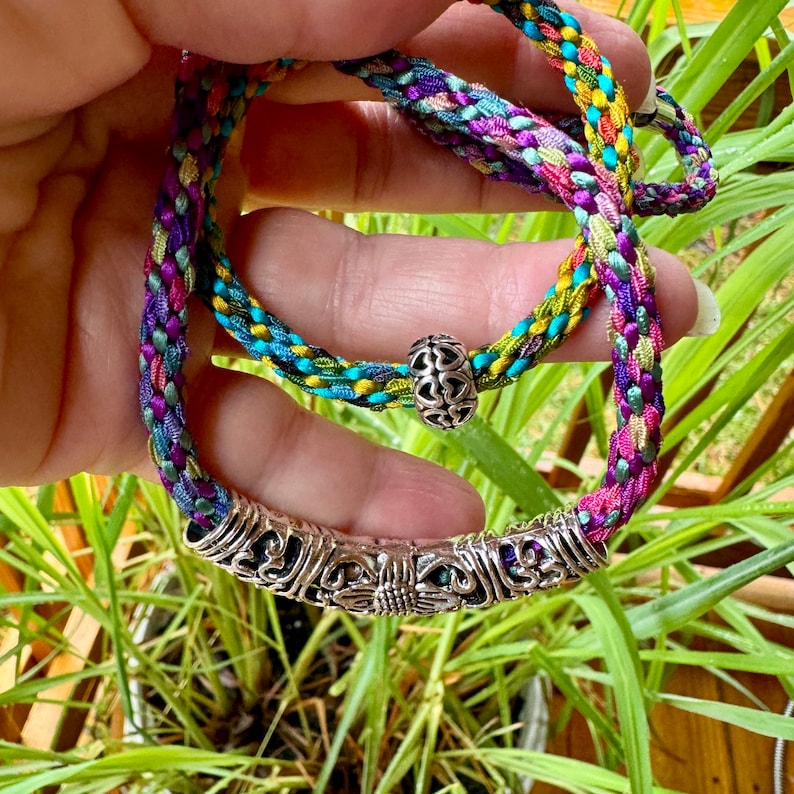 May include: A multi-colored braided cord bracelet featuring a central silver-toned metal tube bead with intricate floral engravings and a smaller silver-toned round bead decorated with a repeating heart pattern along the braided cord.