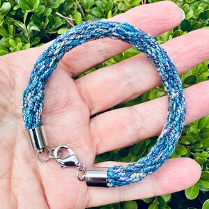 May include: A blue and white braided bracelet with silver metal clasps. The bracelet is made of woven fabric and has a unique, textured look.