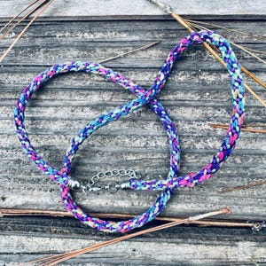 May include: A braided necklace made of multi-colored fabric cord in shades of purple, blue, pink, and teal. The necklace features silver-toned metal end caps and a silver-toned metal lobster claw clasp with an adjustable chain link extension.