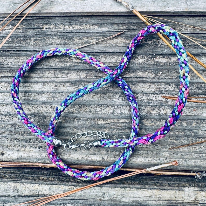 May include: A braided necklace made of multi-colored fabric cord in shades of purple, blue, pink, and teal. The necklace features silver-toned metal end caps and a silver-toned metal lobster claw clasp with an adjustable chain link extension.