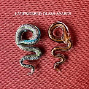 May include: Two lampworked glass snakes with the text LAMPWORKED GLASS SNAKES. One snake is clear with green and blue glittery internal details, while the other is clear with gold glittery internal details. Both are coiled in a serpentine shape.