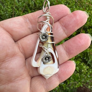 May include: A pendant featuring a conical seashell wrapped in silver-toned wire. The wire forms a loop at the top and includes two small, detailed silver-toned metal flower charms attached to the front of the shell, which has white and brown tones.