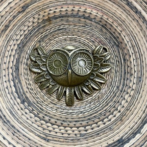 May include: A gold-toned metal owl charm with detailed wings and large eyes. The owl is on a woven background.