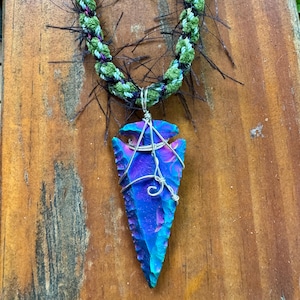 May include: A necklace featuring a vibrant, iridescent blue and purple arrowhead pendant wrapped in silver-toned wire. The pendant is suspended from a braided cord made of green and black fibers with thin, wispy black strands extending outward.