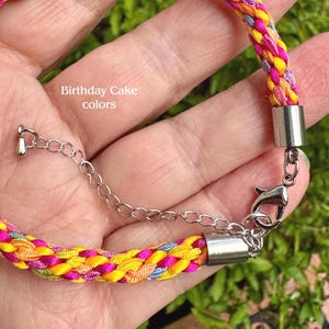 May include: A braided bracelet featuring vibrant yellow, orange, pink, and light blue threads, labeled Birthday Cake colors. The bracelet includes silver-toned metal end caps, a lobster claw clasp, and an adjustable chain link extension.