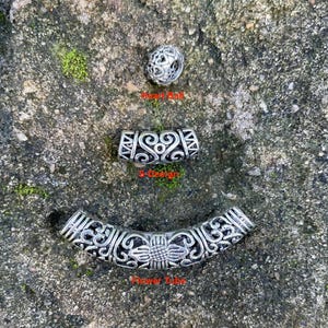 May include: Three silver-toned metal beads featuring intricate cut-out patterns. The top bead is a spherical Heart Ball, the middle is a curved S-Design tube, and the bottom is a long, curved Flower Tube with a central sunflower motif.