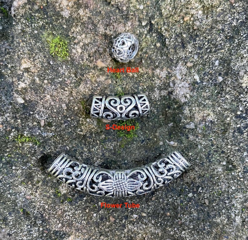 May include: Three silver-toned metal beads featuring intricate cut-out patterns. The top bead is a spherical Heart Ball, the middle is a curved S-Design tube, and the bottom is a long, curved Flower Tube with a central sunflower motif.