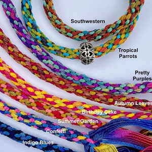 May include: Multiple braided fabric cords in various color combinations labeled Southwestern, Tropical Parrots, Pretty Purples, Autumn Leaves, Birthday Cake, Summer Garden, Confetti, and Indigo Blues. One cord features a silver-toned heart bead.