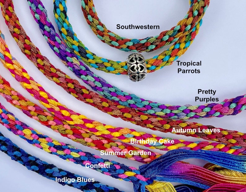 May include: Multiple braided fabric cords in various color combinations labeled Southwestern, Tropical Parrots, Pretty Purples, Autumn Leaves, Birthday Cake, Summer Garden, Confetti, and Indigo Blues. One cord features a silver-toned heart bead.