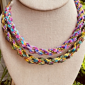 May include: A multi-strand necklace featuring braided yarn in vibrant shades of pink, purple, blue, yellow, and green. The textured, hand-crafted accessory consists of two distinct twisted loops that drape in layers, showcasing a colorful mix of fibers.