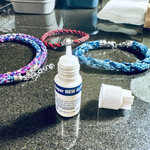 May include: A clear bottle of Super New Glue with a blue and white label. The bottle is sitting on a gray surface with a blue and white braided cord and a red and black braided cord. The bottle cap is off and sitting next to the bottle.