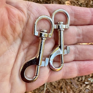 May include: Two shiny silver-colored swivel snap hooks. Each hook has a D-ring at the top and a spring-loaded clasp at the bottom. The hooks are designed for attaching to leashes, straps, or other items.