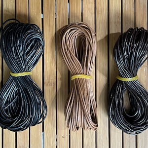 May include: Three spools of leather cord in black, brown, and dark brown. The cords are wrapped around themselves and tied with a yellow rubber band.