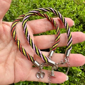 May include: Two braided cord bracelets featuring a repeating pattern of black, yellow, orange, and pink threads. Each bracelet is finished with silver-toned metal end caps, a lobster claw clasp, an adjustable chain, and a small silver-toned charm.
