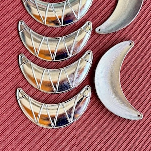 May include: Silver-toned metal crescent moon charms with a textured surface and small holes for stringing. The charms have a geometric design and are arranged on a red background. Ideal for jewelry making or craft projects.