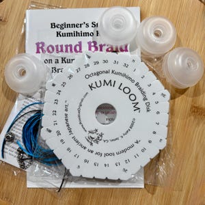 May include: A white octagonal Kumi Loom braiding disk with numbered slots from 1 to 32, accompanied by four translucent plastic bobbins, a bundle of blue and black cord, and an instructional booklet titled Beginner's Kumihimo Round Braid.