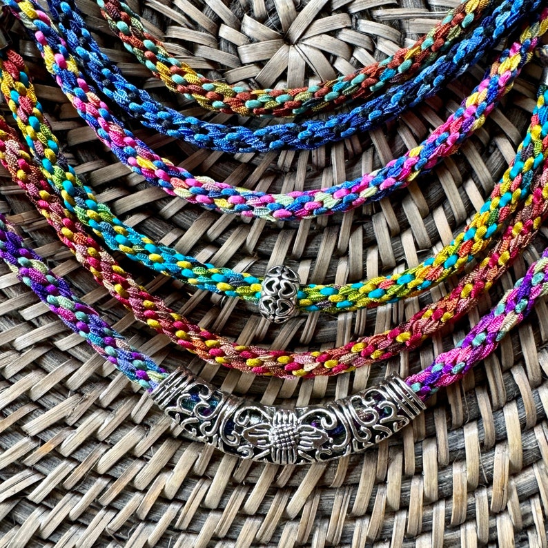 May include: A multi-layered necklace featuring several braided cords in vibrant shades of blue, pink, yellow, and green. The necklace includes a small silver-toned bead with a heart pattern and a larger, curved silver-toned metal focal piece.