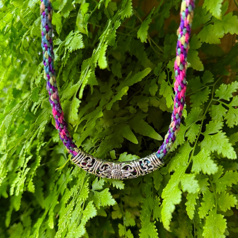 May include: A braided necklace featuring a multi-colored cord in shades of pink, purple, blue, and yellow. The cord is threaded through a curved, silver-toned metal tube bead with an intricate, embossed floral and scrollwork pattern.