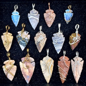 May include: A collection of handcrafted arrowhead pendants. Each pendant features a unique stone, including blue, gray, and brown varieties, intricately wrapped with silver, gold, and copper wire. The pendants are displayed against a dark background.