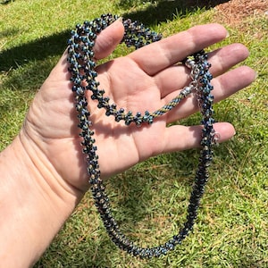 May include: A beaded necklace with a dark blue and green iridescent color scheme. The necklace is held in a hand, showcasing its intricate design and the way it catches the light. The clasp is silver.