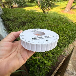 May include: A white foam mini Kumihimo disk with a circular center hole, featuring black printed text that reads Bear Smith Double-Density, mini Kumihimo Disk, and Create your own braided cord, along with numbered slots from 1 to 32 around the edge.