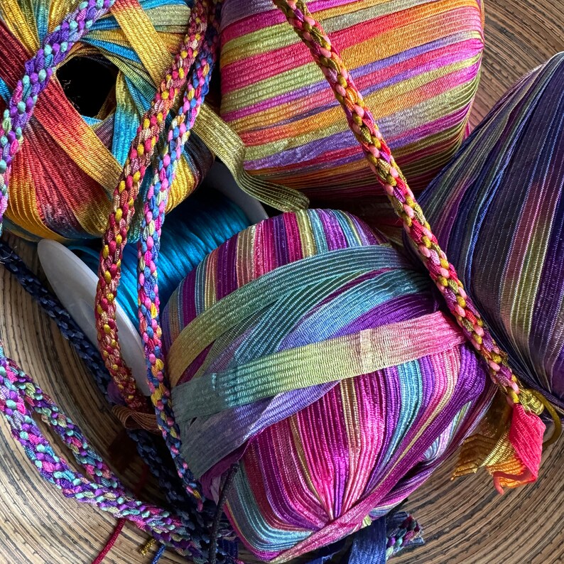 May include: Several spools of vibrant, multi-colored ribbon featuring metallic threads and striped patterns in shades of pink, purple, blue, gold, and green. A braided trim in matching variegated colors winds across the spools of flat ribbon.