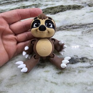 May include: A brown and tan sloth figurine with white claws and a smiling face. The figurine is made of plastic and has articulated limbs.