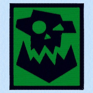 Puede incluir: Un parche bordado negro y verde con un diseño de calavera y dientes. La calavera está delineada en blanco y tiene un ojo verde. Los dientes son irregulares y puntiagudos. El parche está rodeado por un borde negro.