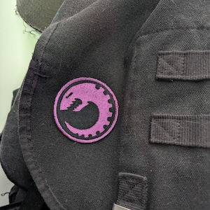 Könnte beinhalten: Eine schwarze Umhängetasche mit einem gestickten, violetten Aufnäher, der einen stilisierten Drachen mit einem Zahnrad-Design zeigt.