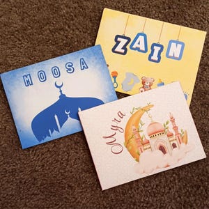 以下が含まれることがあります： 異なるデザインのパーソナライズされたカードが3枚。最初のカードは、白いモスクのシルエットと白い文字で「MOOSA」と書かれた青いカード。2枚目は、青い文字で「ZAIN」と書かれた黄色のカード。3枚目は、三日月、モスク、そして茶色の文字で「MYRA」と書かれたピンクのカードです。