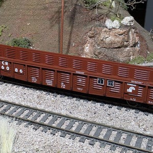 May include: A brown model train gondola car featuring the text CR 57 60 86 on its side. The car has a slatted design, white lettering, and a small blue and white logo. It is positioned on a miniature railway track with gravel ballast.