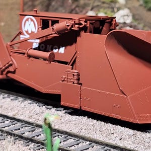 May include: A brown model railroad snowplow car featuring a large front plow blade, side wings, and the white text MONON with a circular logo on the side. The train car is positioned on a set of metal tracks with detailed mechanical components.