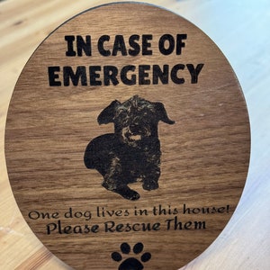 May include: A wooden sign with the text "In Case of Emergency" and a black and white photo of a dog. The sign also says "One dog lives in this house! Please Rescue Them" and has a paw print.