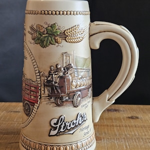 May include: Cream-colored ceramic beer stein with a large handle. The stein features embossed images of hops, wheat, and a vintage beer delivery truck. The text "Stroh's Heritage Series V" is displayed prominently on the front.