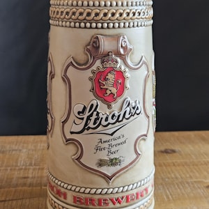 May include: A beige ceramic beer stein with the Stroh's logo, featuring a red shield with a lion and crown. The stein has a decorative border and the words "America's Fire-Brewed Beer".
