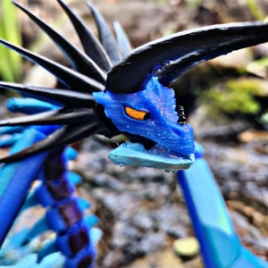 May include: A blue and black dragon figurine with orange eyes. The dragon has a detailed, textured design, with spiky protrusions along its back and head. The figurine is set against a blurred natural background.