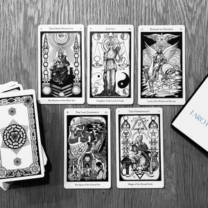 May include: A spread of tarot cards on a wooden table, including The High Priestess, Justice, Knight of Swords, The Last Judgement, and The Hierophant. The cards are black and white with intricate designs. A tarot reading guide book is also visible.