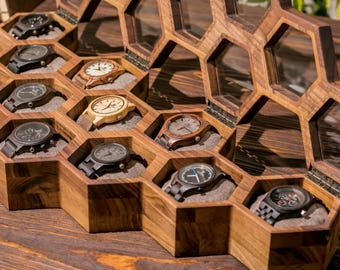 May include: A wooden watch display case featuring a honeycomb-inspired design with multiple hexagonal compartments. The case has a hinged lid with glass panels and contains several wristwatches with wooden and metal bands resting on gray cushions.