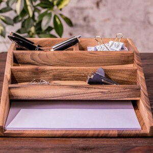 May include: A wooden desk organizer with four compartments. The top compartment has two black pens and a pair of silver paper clips. The second compartment has a silver paper clip. The third compartment has a silver paper clip and a black stapler. The bottom compartment has a white piece of paper.