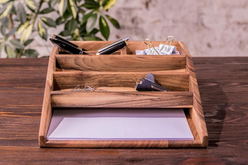 May include: A wooden desk organizer with four compartments. The top compartment has two black pens and a pair of silver paper clips. The second compartment has a silver paper clip. The third compartment has a silver paper clip and a black stapler. The bottom compartment has a white piece of paper.