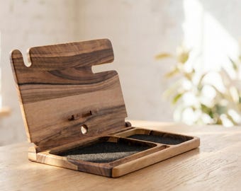 May include: A wooden desk organizer with a natural wood grain finish. The organizer has a slot for a phone and two compartments with black felt lining. It is open and angled, resting on a light-colored wooden surface.