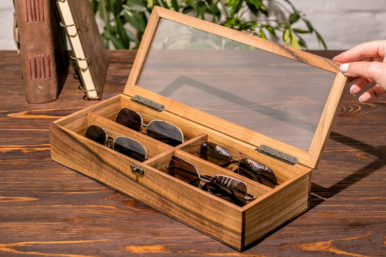 Puede incluir: Una caja de madera con tapa de vidrio para guardar gafas de sol. La caja tiene cinco compartimentos y est&aacute; llena de gafas de sol.