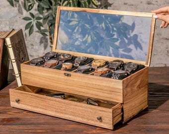 May include: A rectangular wooden watch storage box with a hinged glass lid and a pull-out drawer. The interior features ten individual slots lined with dark fabric, each containing a wristwatch. The box has a natural light brown wood grain finish.