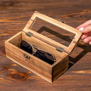 Puede incluir: Una caja de madera con tapa de vidrio, abierta para revelar un par de gafas de sol en su interior. La caja est&aacute; hecha de madera marr&oacute;n claro con un acabado natural. Las gafas de sol son negras con un marco dorado.